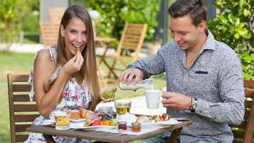 A couple dining outdoors at a wooden table with a meal, including bread, fruits, and a beverage.