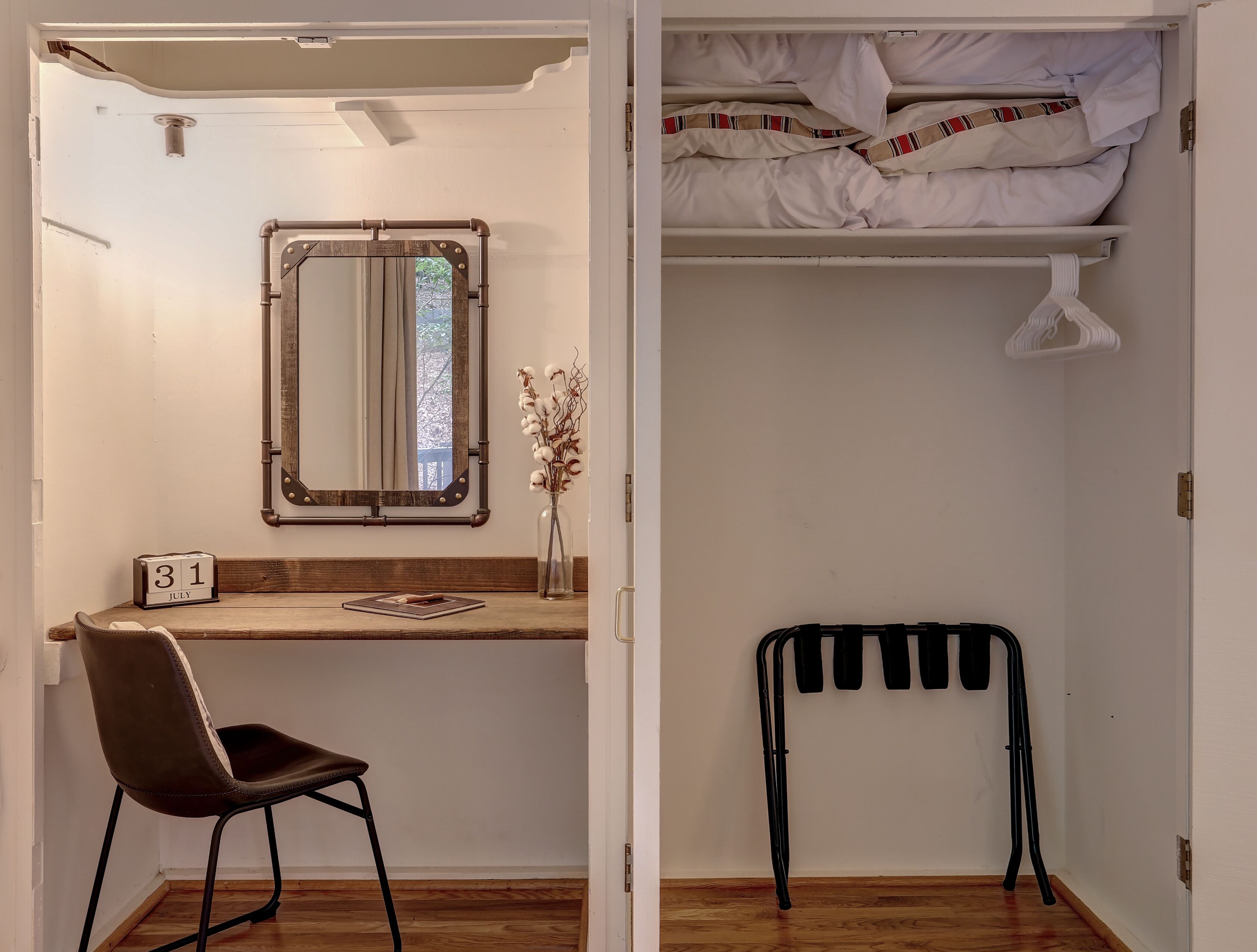 A compact room with a wooden desk, a chair, a mirror, a date calendar, a hanging chair, and a bed with neatly folded blankets.