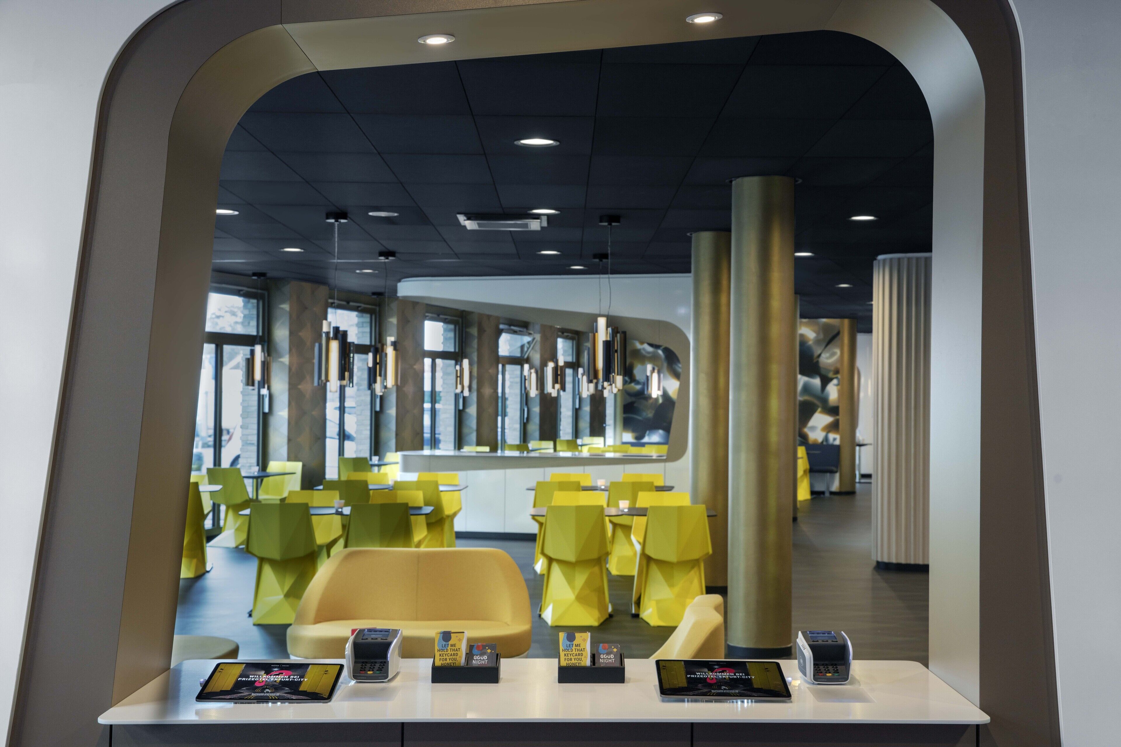 Un intérieur moderne avec des chaises et des tables jaunes, un comptoir d’accueil et une grande fenêtre en arrière-plan.