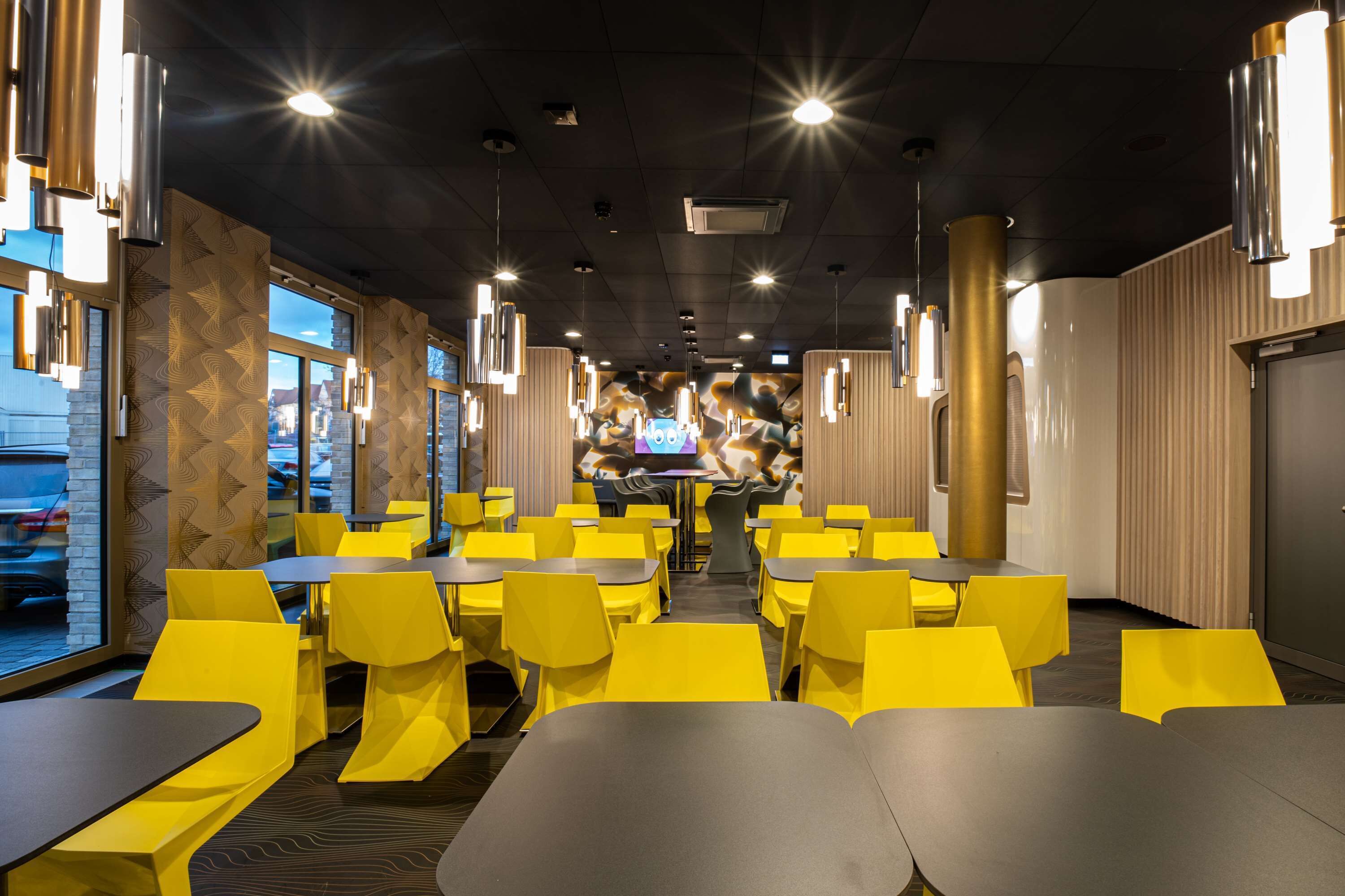A restaurant with yellow chairs and tables, a counter with a person at the back, and large windows on the left.