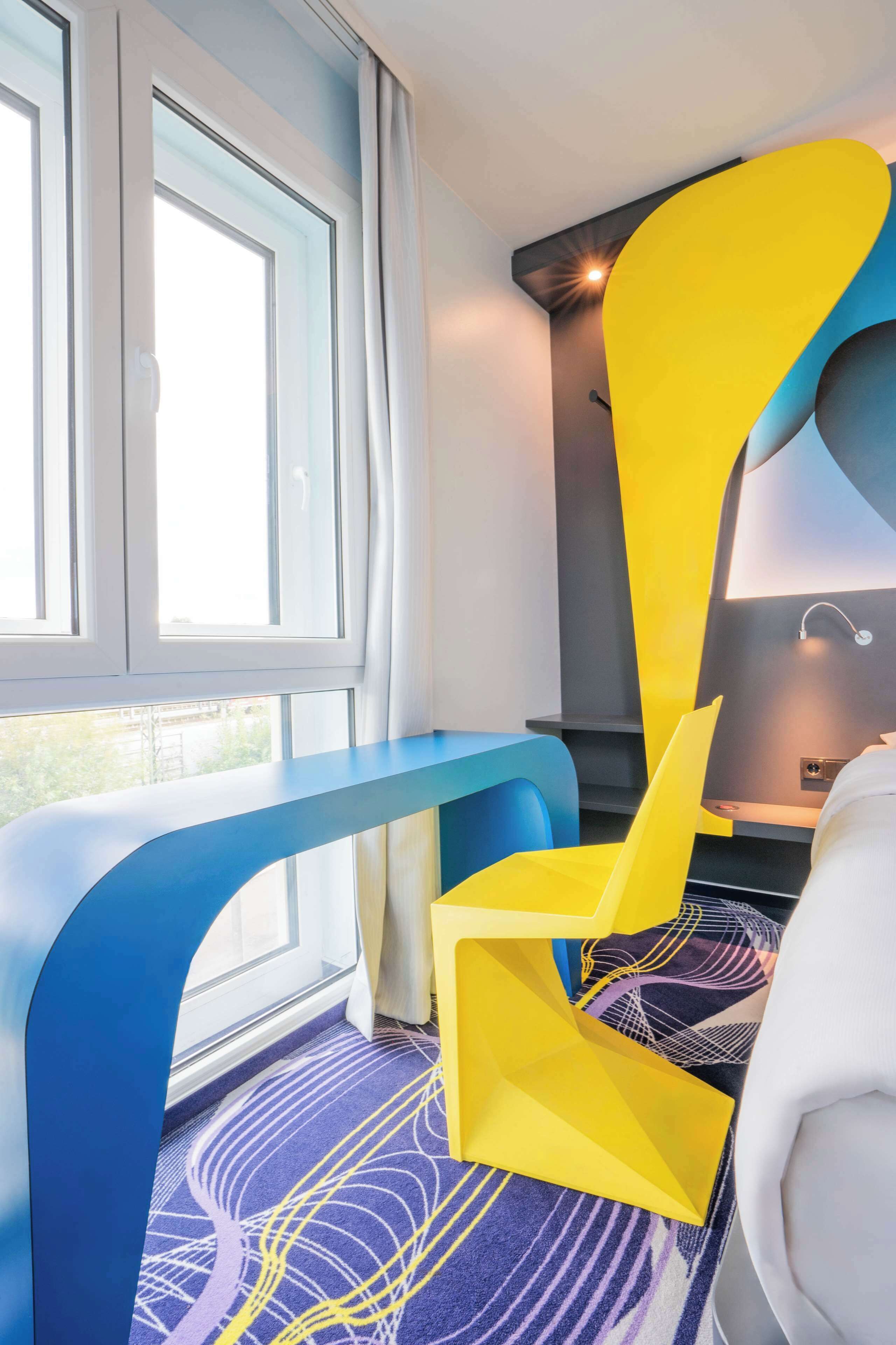 A modern room with a large window, a blue bench, a yellow chair, and a patterned carpet.