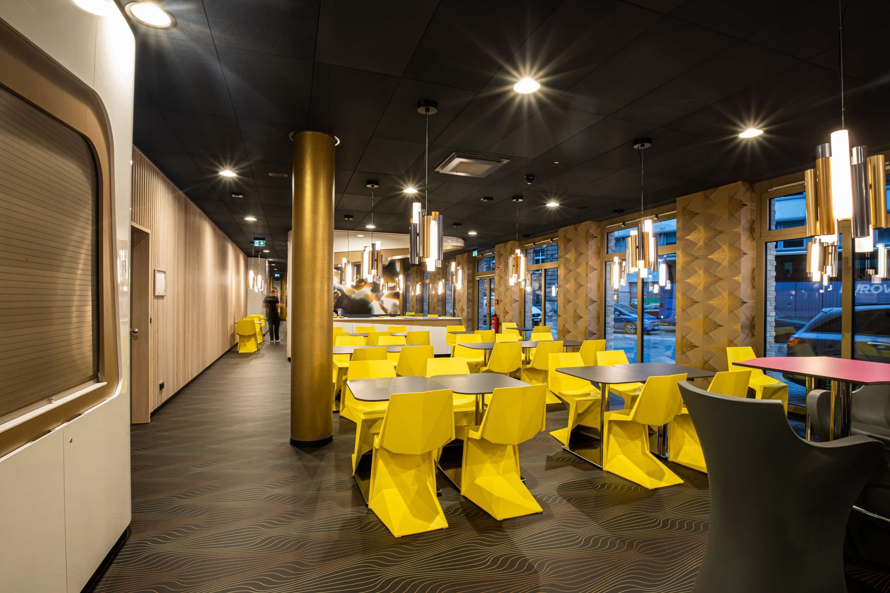 Een moderne eetruimte met gele stoelen en tafels, grote ramen en een vloer met een patroon.