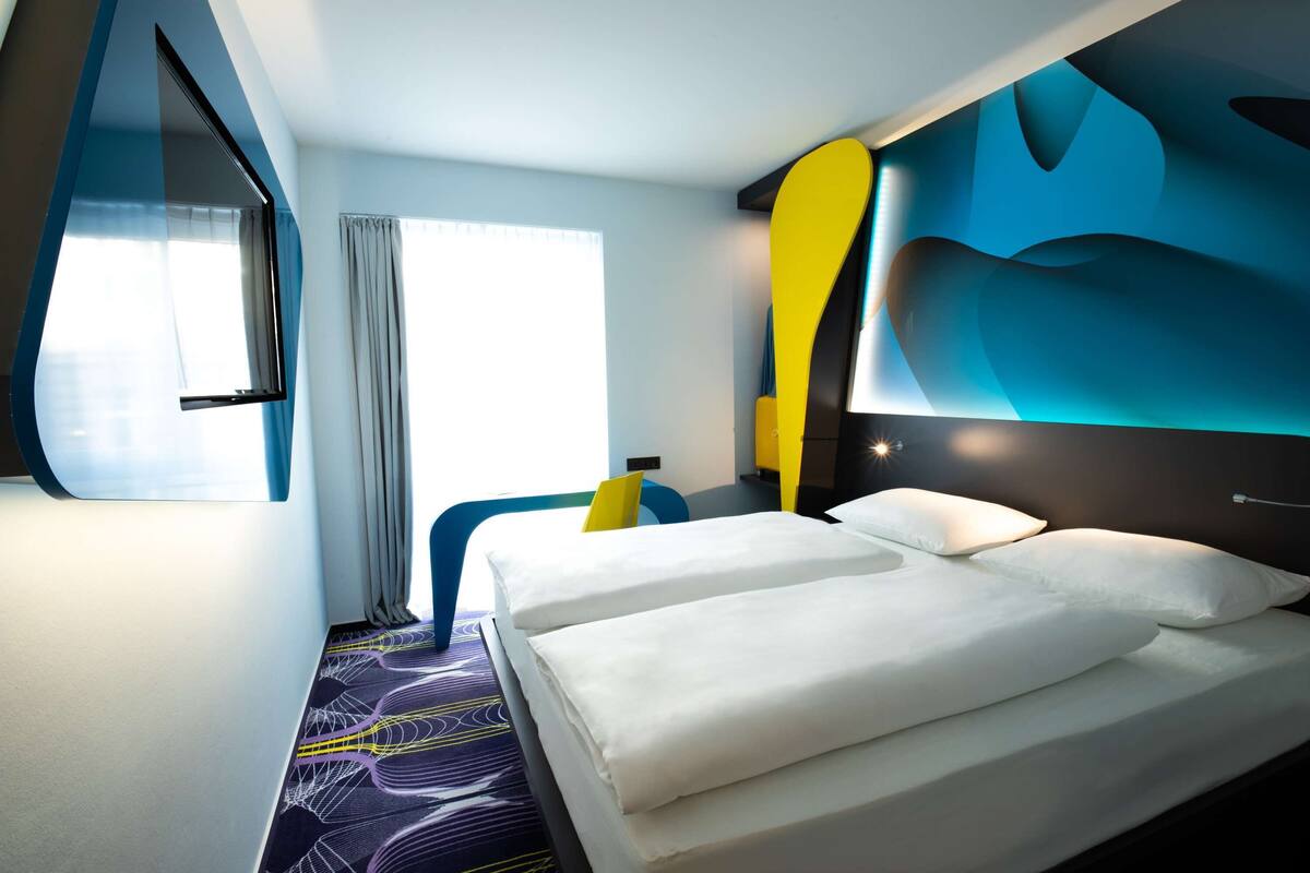 A modern hotel room with a large bed, a desk, and a window with curtains.