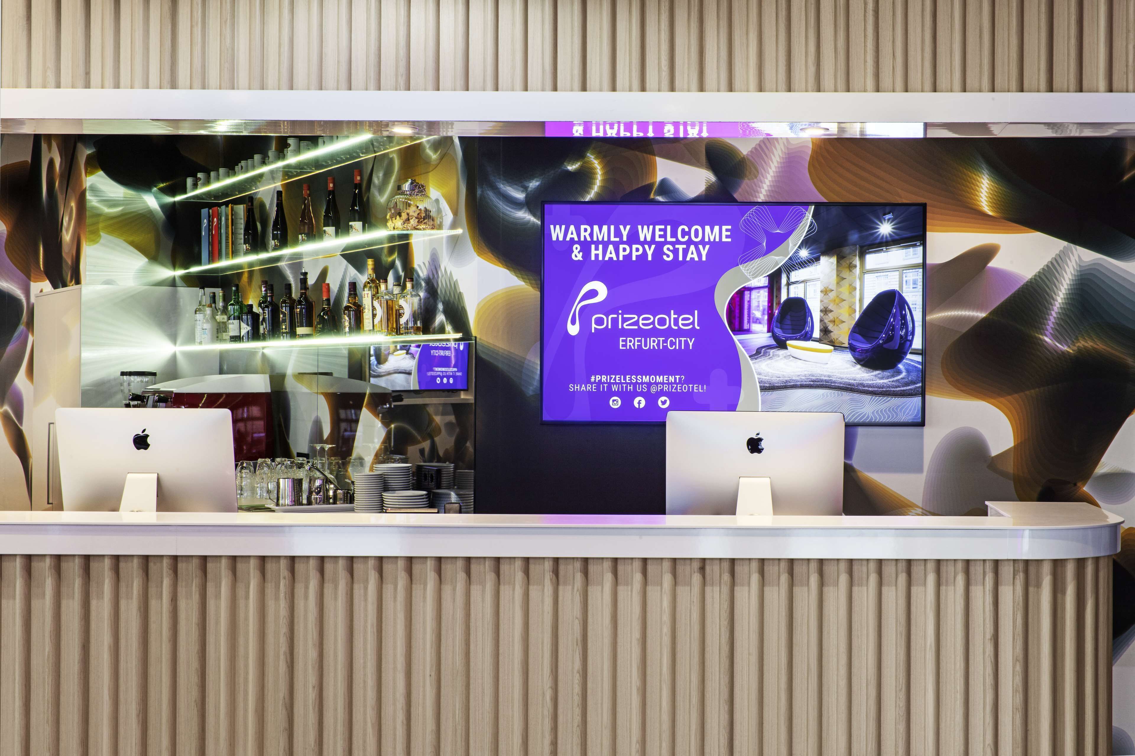 A hotel reception desk with a digital screen displaying a welcome message, a bar area with various bottles, and a seating area with chairs.