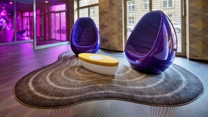 Un interior moderno con dos sillas moradas de forma ovoide sobre una alfombra estampada, un pequeño otoman blanco y amarillo, y ventanas amplias con vistas a edificios.