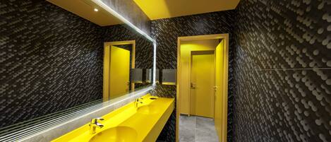 Un baño moderno con un lavabo y espejo amarillo, paredes de un color oscuro con diseño, y una puerta.