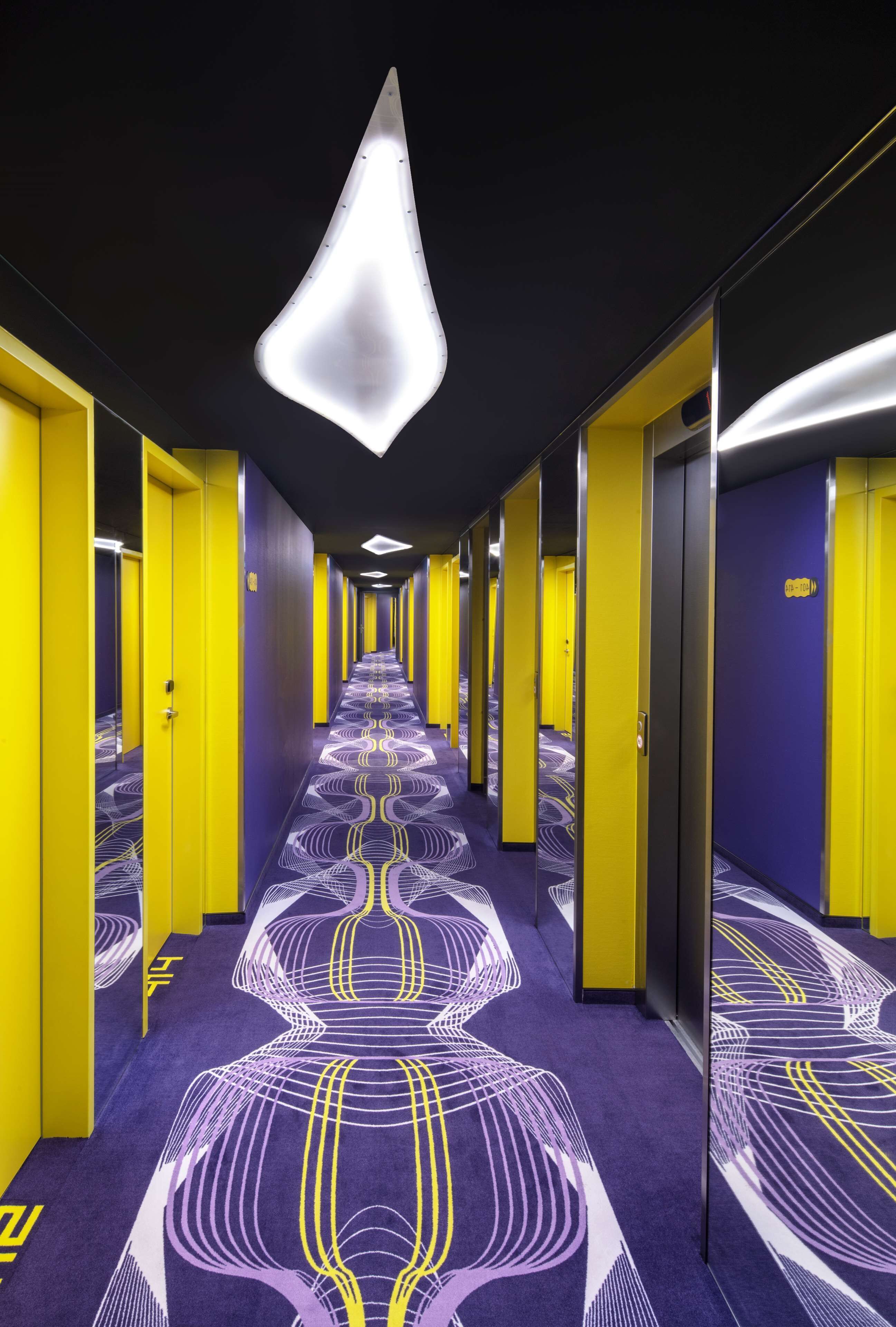 Un couloir d’hôtel aux murs recouverts de miroirs, aux portes jaunes et au tapis à motifs.
