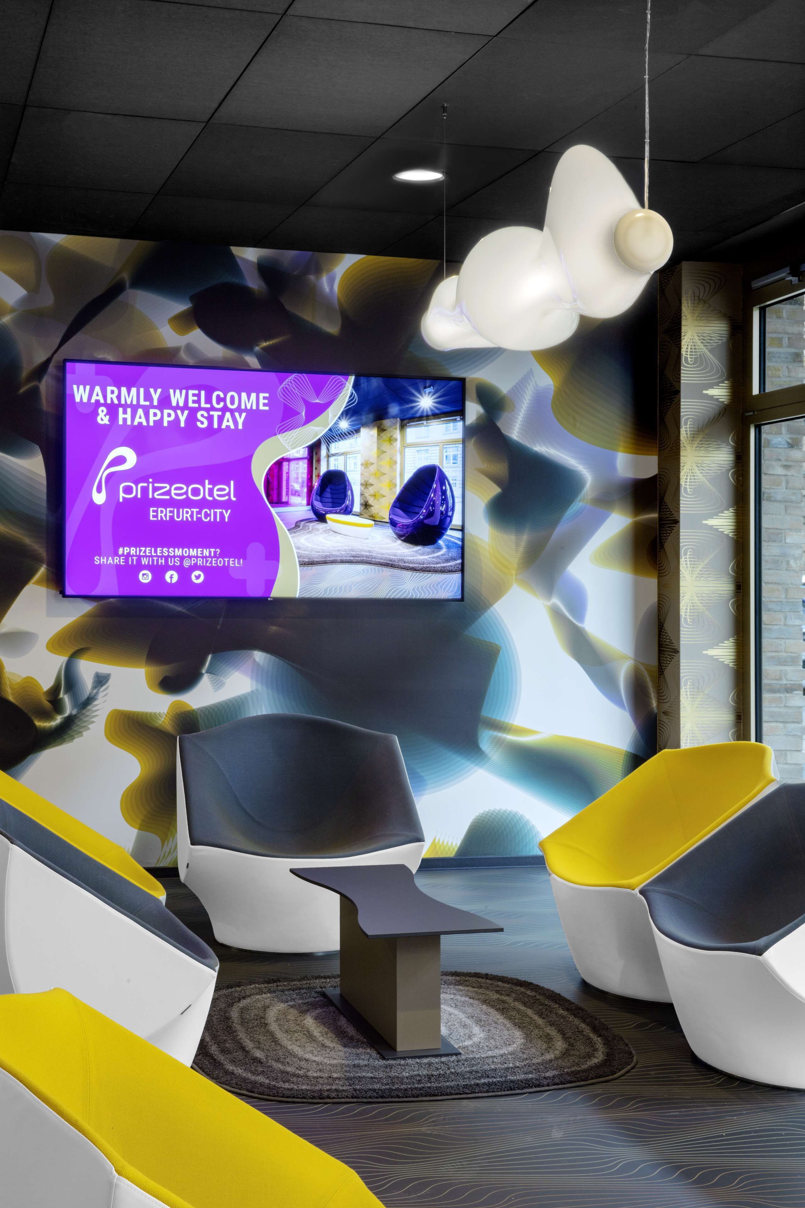 A modern hotel lobby with a large TV screen displaying a welcome message, unique seating arrangements, and a patterned wall design.
