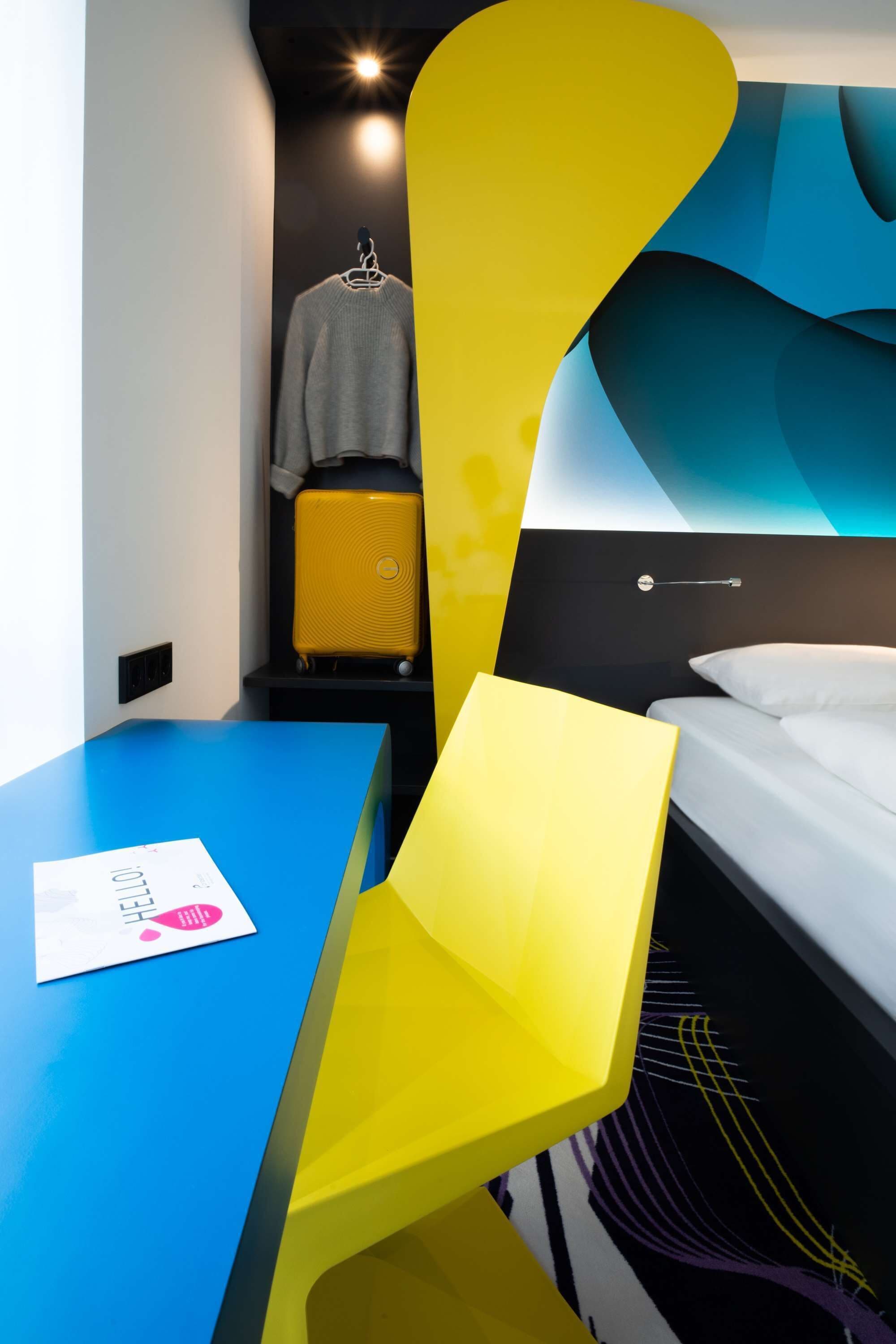 Ein modernes Hotelzimmer mit einem blauen Schreibtisch, einem Bett mit weißen Leinen und einem gelben Koffer auf einem Regal.