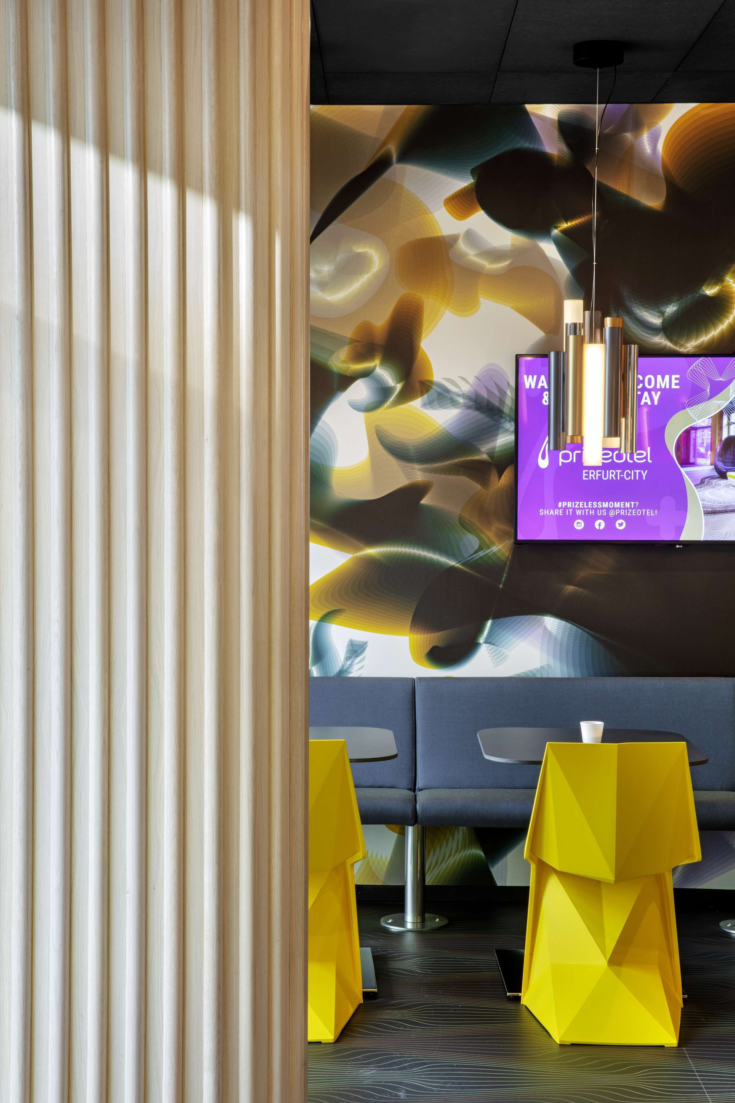 En moderne hotellobby med et stort digitalt skærm, der viser en velkomstbesked, en moderne lampe og gule, geometriske siddepladser.