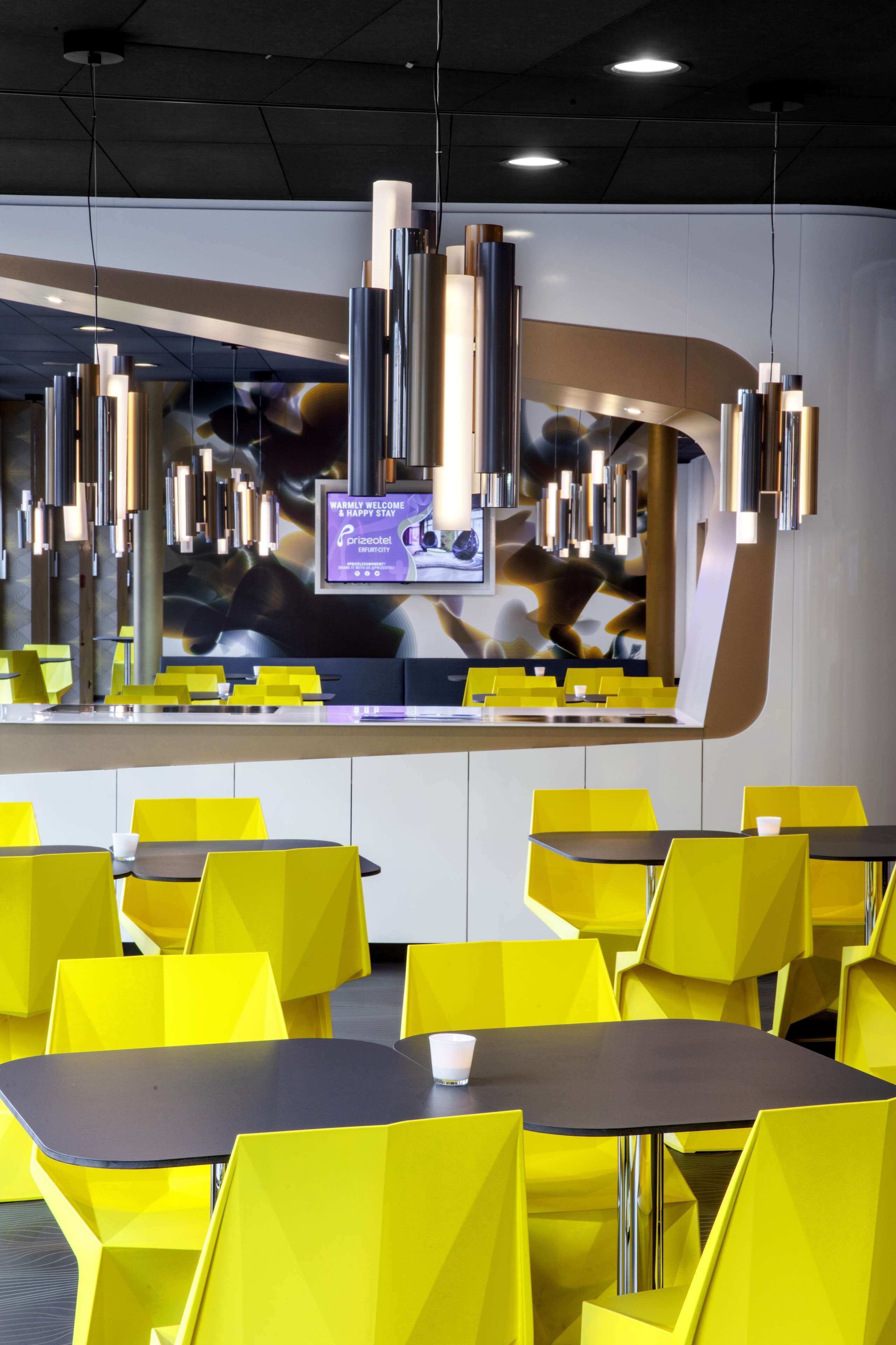 Un restaurant moderne avec des chaises et des tables jaunes, un écran numérique affichant un message de bienvenue et des luminaires au design contemporain.