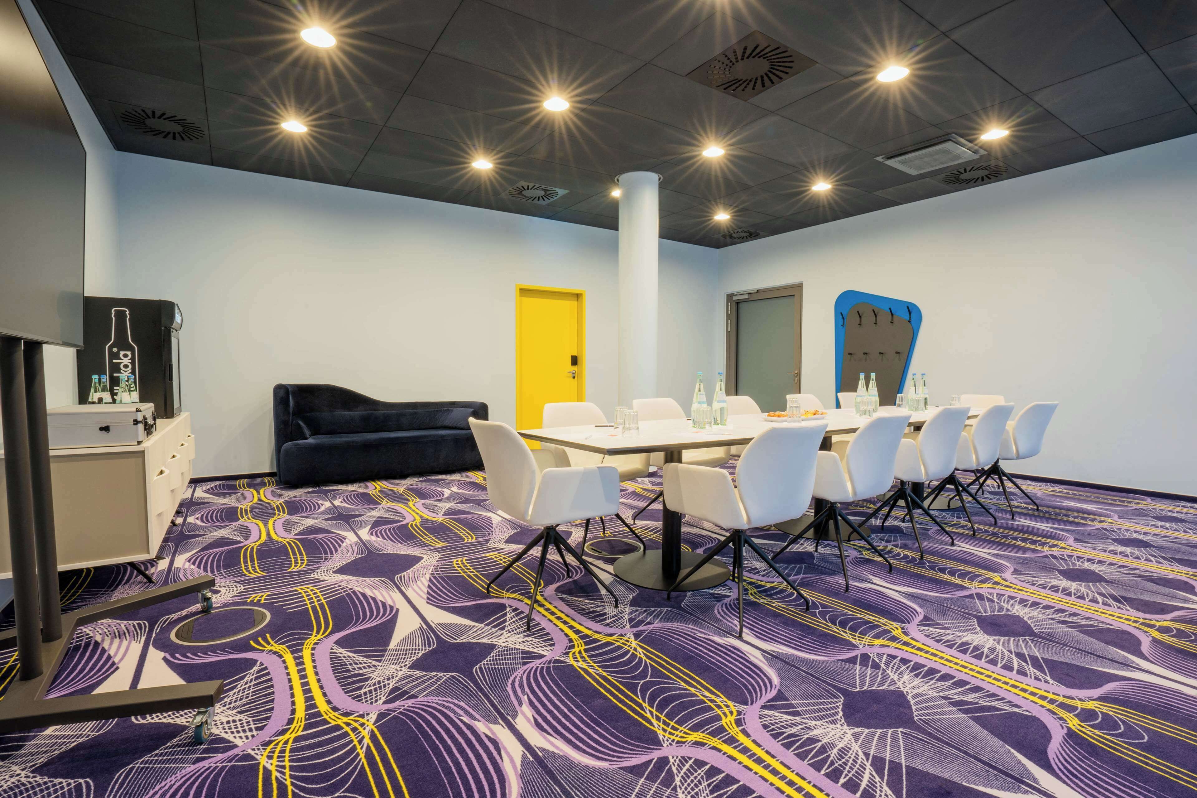Een vergaderruimte met een paars patroon tapijt, een witte tafel met stoelen, een zwarte bank en een gele deur.