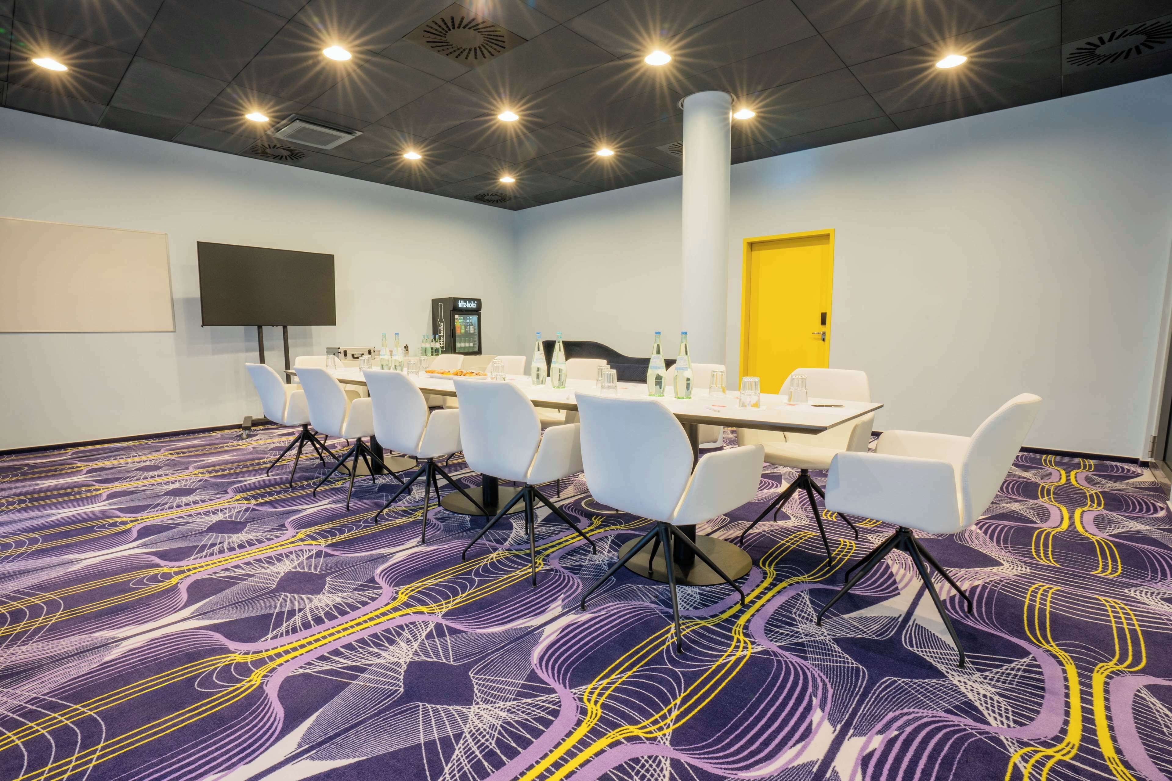 Un salón de conferencias con una mesa grande, sillas blancas, una pantalla de proyector y una puerta amarilla.