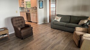 Ein Wohnzimmer mit einem braunen Sessel, einem grünen Sofa und einem Korbstuhl. Durch eine offene Tür ist eine Küche erkennbar.