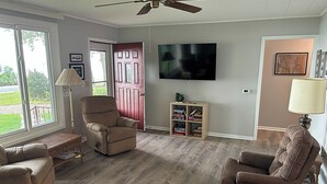 Ein Wohnzimmer mit zwei beigen Sesseln, Holzboden, Deckenventilator, Fernseher und Bücherregal.
