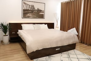 A modern bedroom with a large bed, a headboard, a bedside table, a potted plant, a wall-mounted artwork, and a window with curtains.