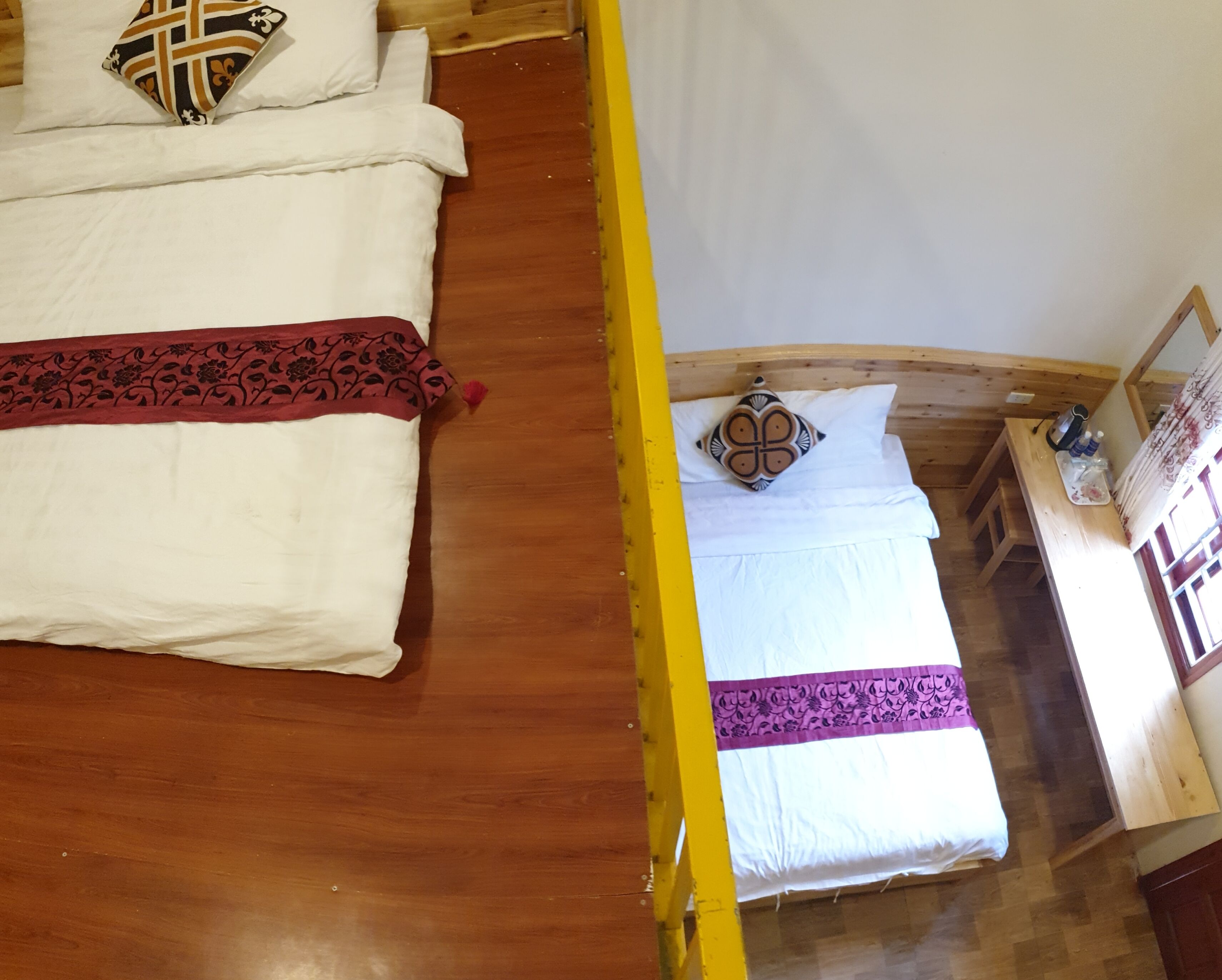 A room with two beds, each with a distinct patterned pillow. The beds are made with white linens and have decorative pillows. A wooden bench runs along the wall, and a small table with a bottle and a tray is visible.