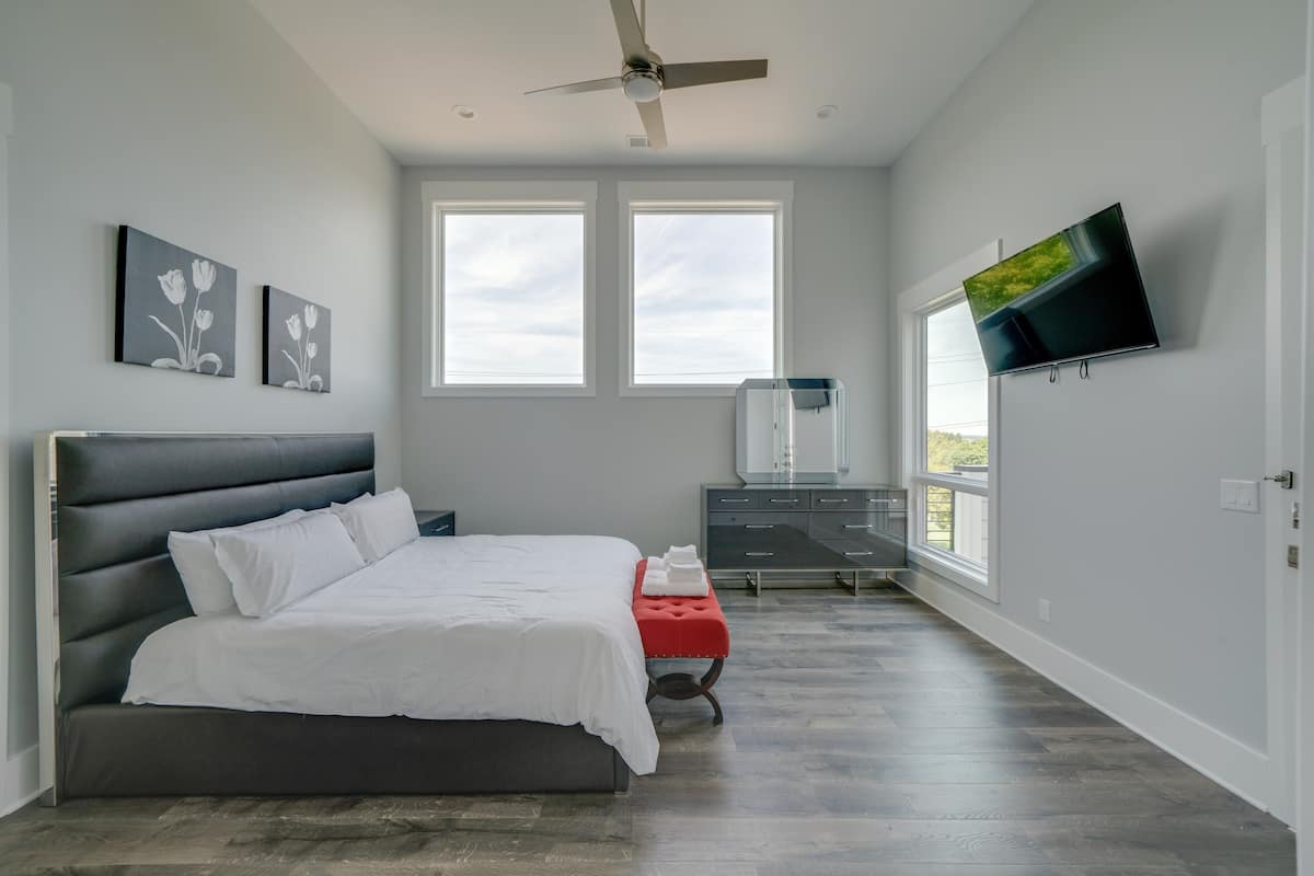 Un dormitorio moderno con una cama grande, un ventilador de techo, dos ventanas, un televisor y un aparador.