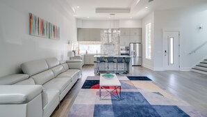 Ein modernes Wohnzimmer mit einer Couch, einem Essbereich mit Stühlen, einer Küche im Hintergrund und einer Treppe.