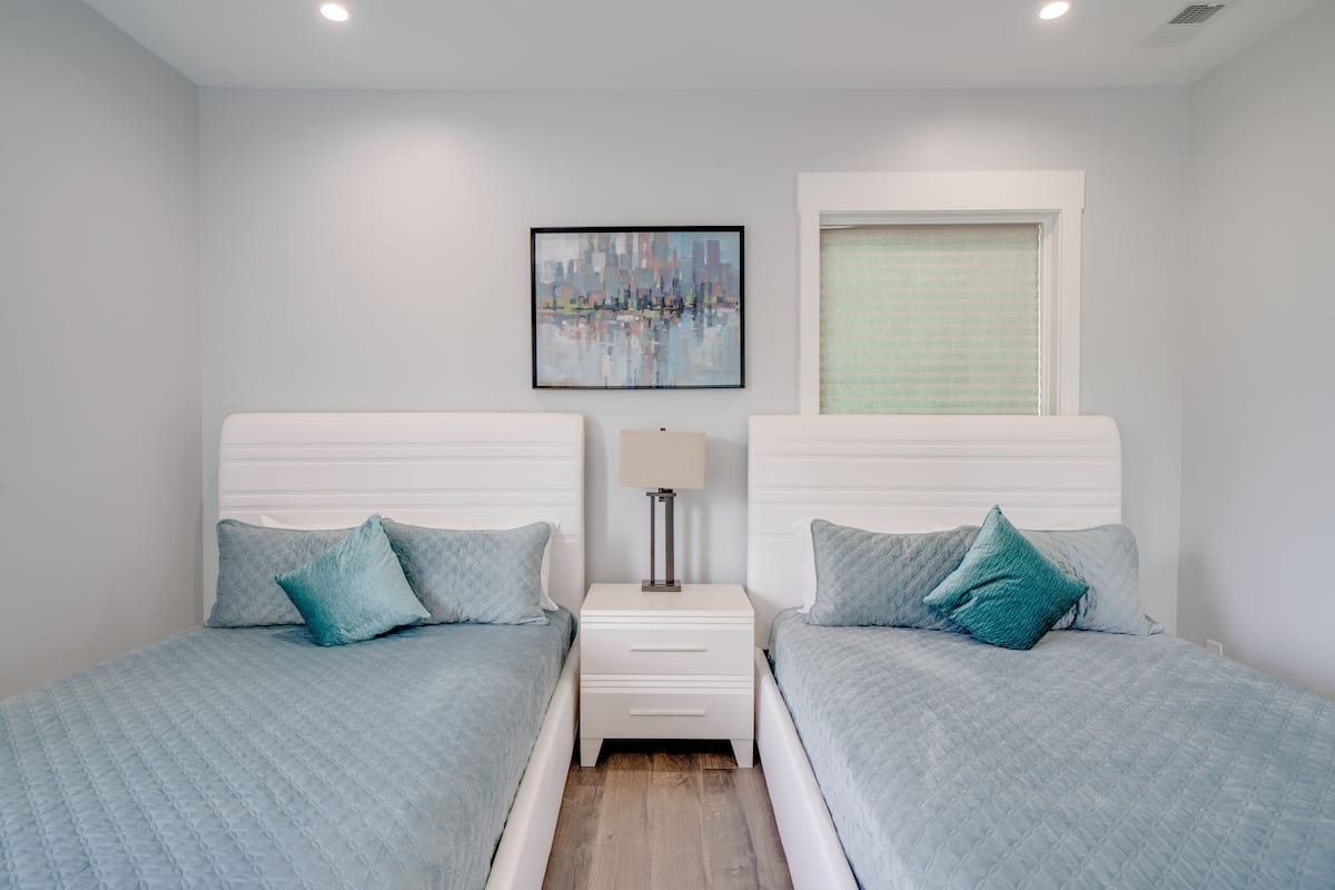 Un dormitorio con dos camas, una mesita de noche, una lámpara, una ventana con persianas y una pintura de paisaje urbano en la pared.