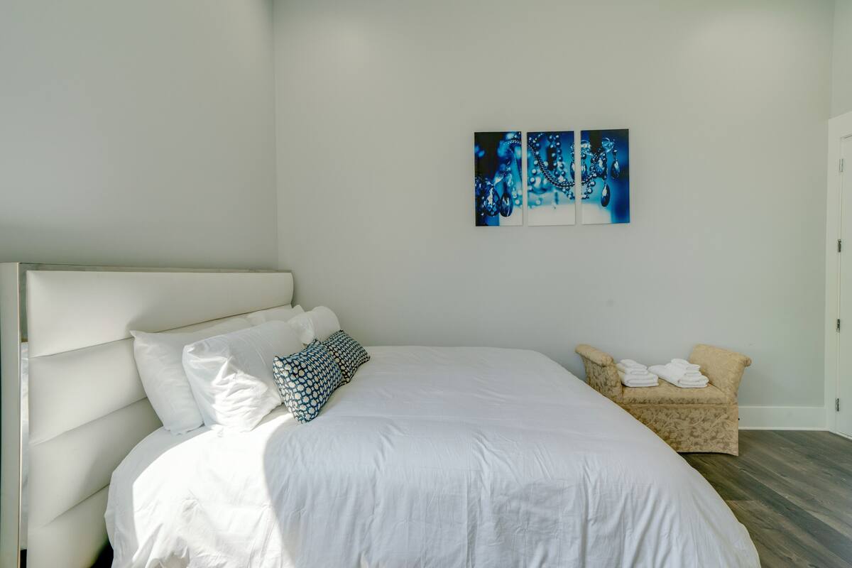 Una cama bien hecha con sábanas blancas y un cojín estampado, un sillón beige con toallas, y arte abstracto en la pared.