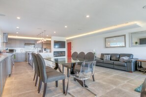 A modern kitchen with grey cabinets, a built-in oven, and a microwave. There is a dining area with a glass table and grey chairs. A grey sofa is in the living area, and a red door is visible. - Beach Front 6000SF  10b/6bth  7 Miles  to SF Moscone Center (Pacifica)
