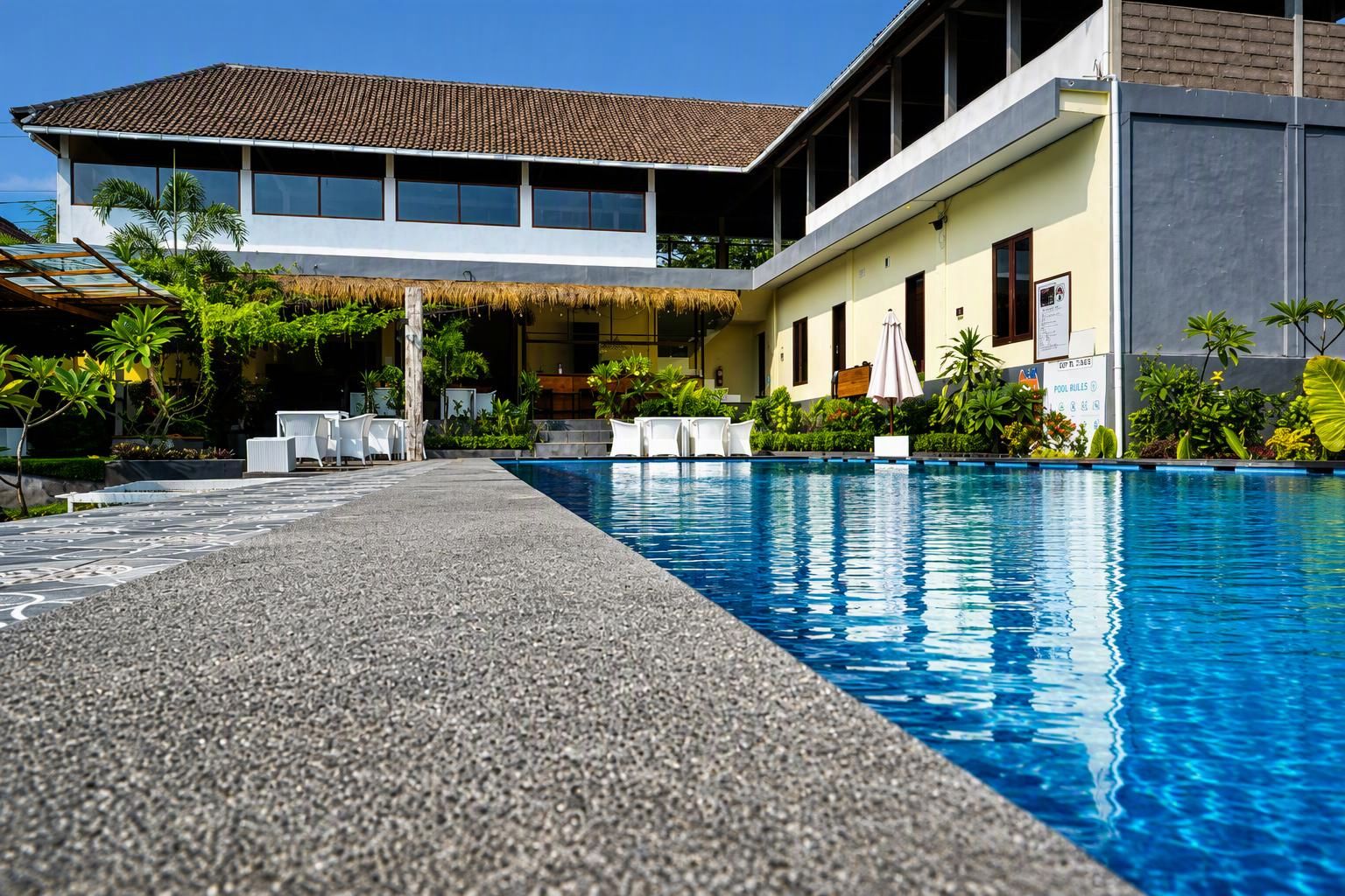 A swimming pool with a clear blue water surface, surrounded by a paved walkway, and adjacent to a building with a tiled roof and a thatched structure.