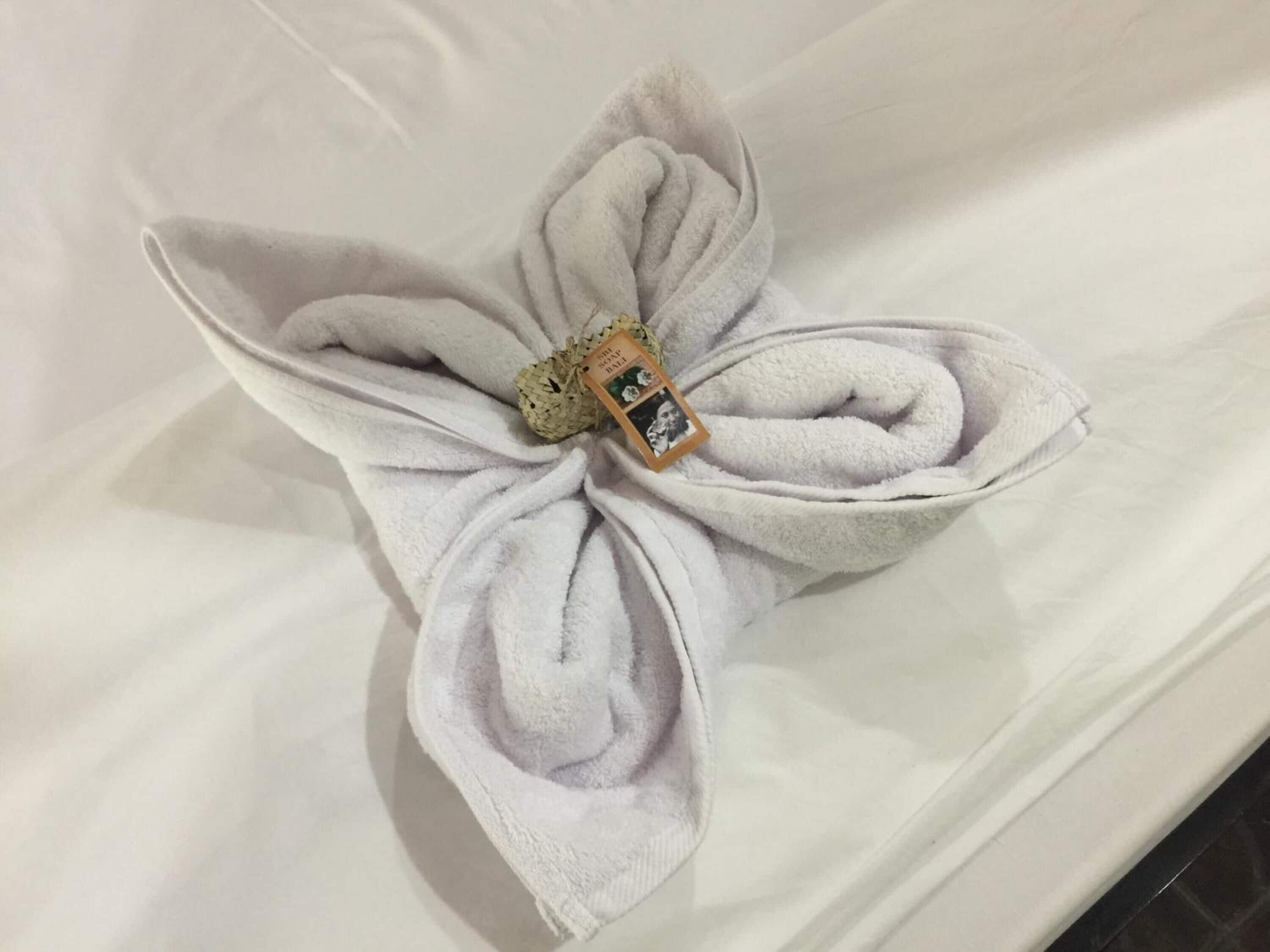 Ein Handtuch, zu einer Blume geformt, liegt auf einer weißen Oberfläche, vermutlich einer Badezimmerablage.