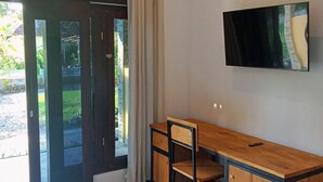 Quarto com escrivaninha de madeira, cadeira, televisão fixada na parede e porta de vidro deslizante para área externa.