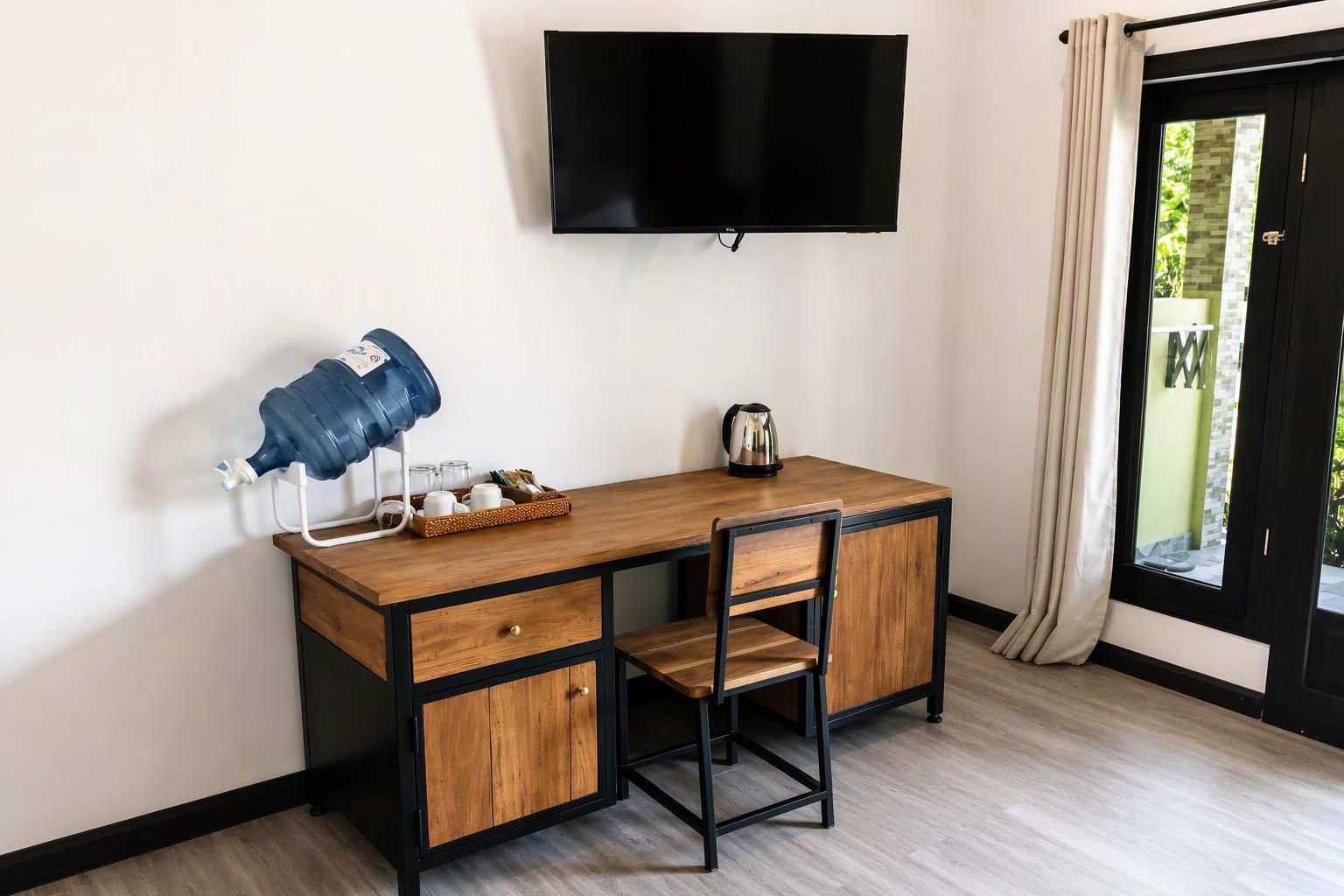 A room with a wooden desk, a chair, a television mounted on the wall, and a window with curtains.