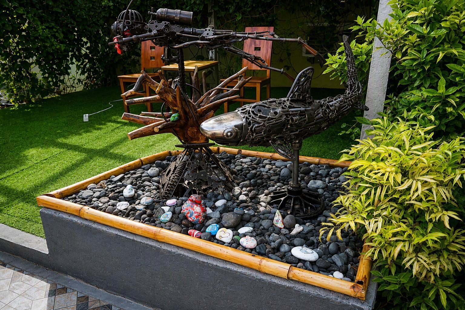 Un jardín con una escultura única de un ave hecha con metal y ramas, rodeado por una jardinera llena de piedras y un pequeño árbol.