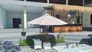 Uma área de estar externa com móveis de vime branco, um telhado de palha e um espaço de bar com assentos de madeira.