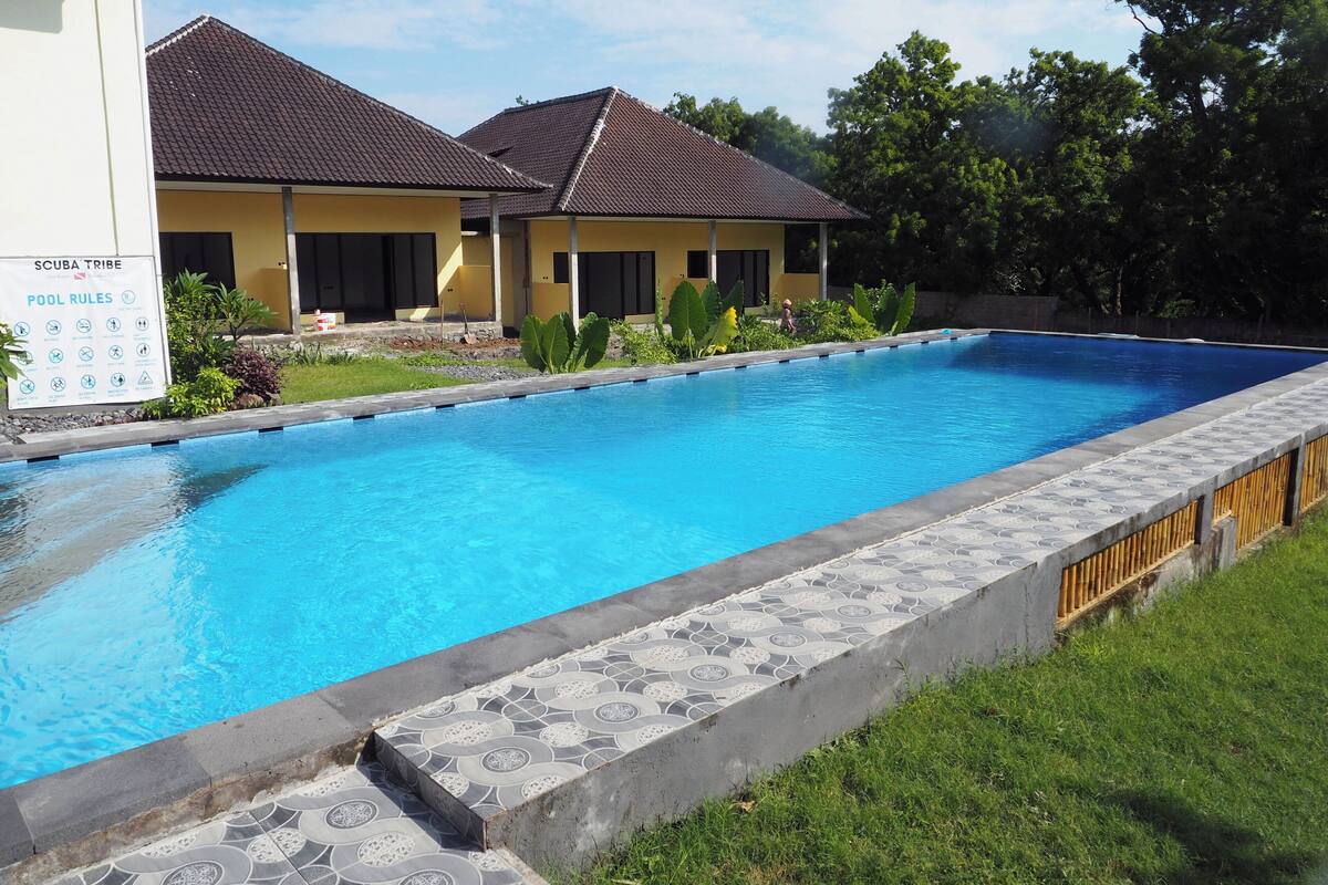 A swimming pool with tiled steps leading into it, surrounded by a grassy area and a building with a tiled roof.