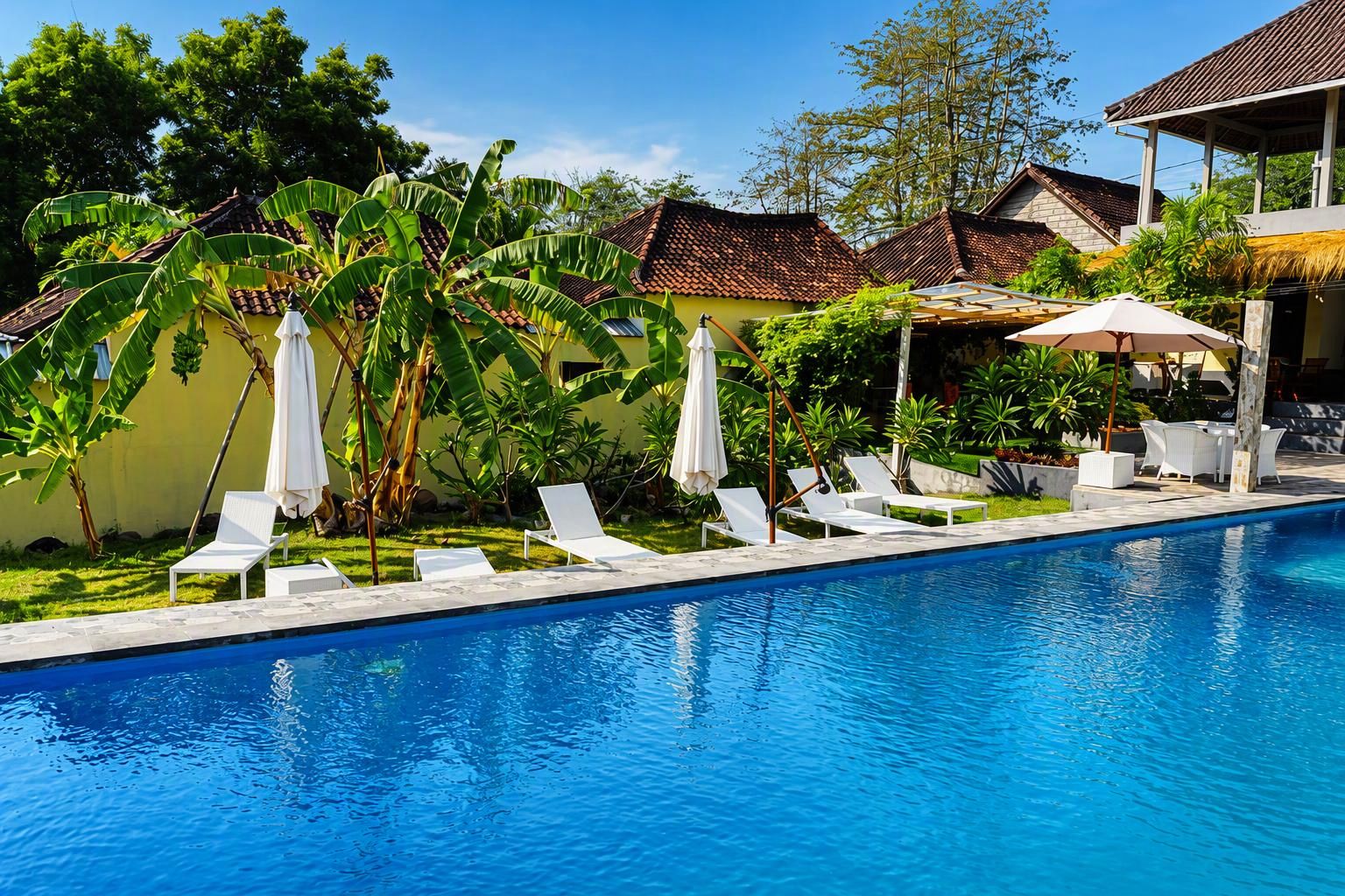 A swimming pool with white lounge chairs and umbrellas, surrounded by tropical vegetation and buildings with tiled roofs.