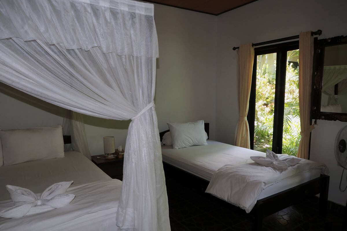 A room with two beds, each with white linens and pillows, a bedside table with a lamp, and a window with curtains.