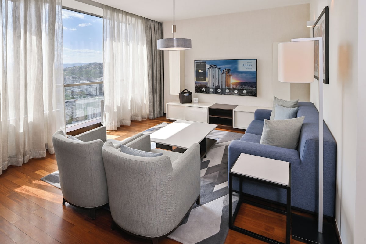 Ein modernes Wohnzimmer mit einer Couch, Sesseln, einem Couchtisch, einem Fernseher und Blick auf die Stadt.