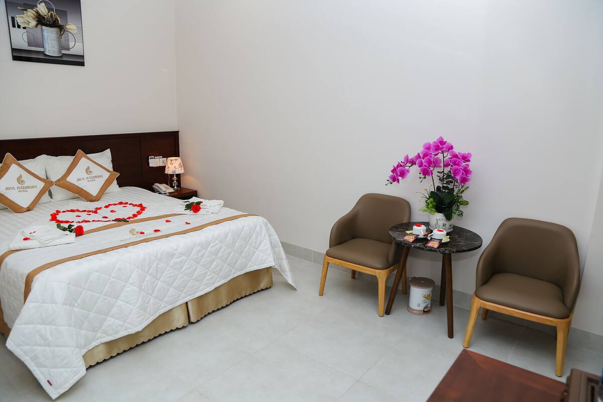 A neatly made bed with a white quilt and decorative pillows, a small round table with a vase of flowers, and two brown chairs in a hotel room.