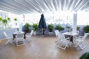A rooftop terrace with white folding chairs and tables, surrounded by potted plants and a large umbrella.
