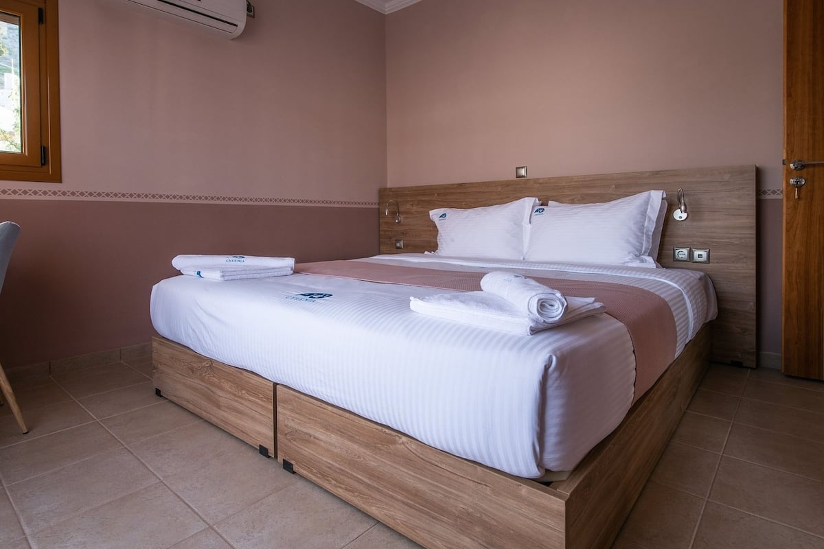 A neatly made bed with white linens and a wooden headboard, a bedside table, and a window with blinds.