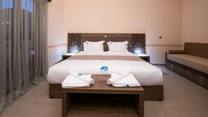 Habitación de hotel con una cama grande, un cabecero de madera, dos mesitas de noche con lámparas, un sofá y una ventana con cortinas traslúcidas.