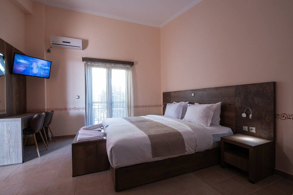 A hotel room with a large bed, a desk with two chairs, a TV, and an air conditioning unit.