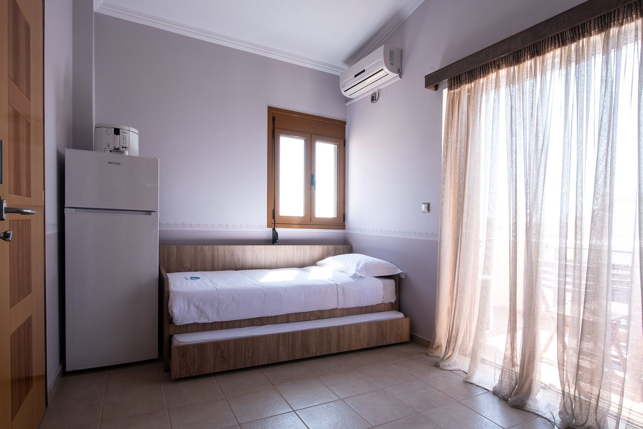 Una habitación pequeña con cama, ventana, puerta y aire acondicionado.