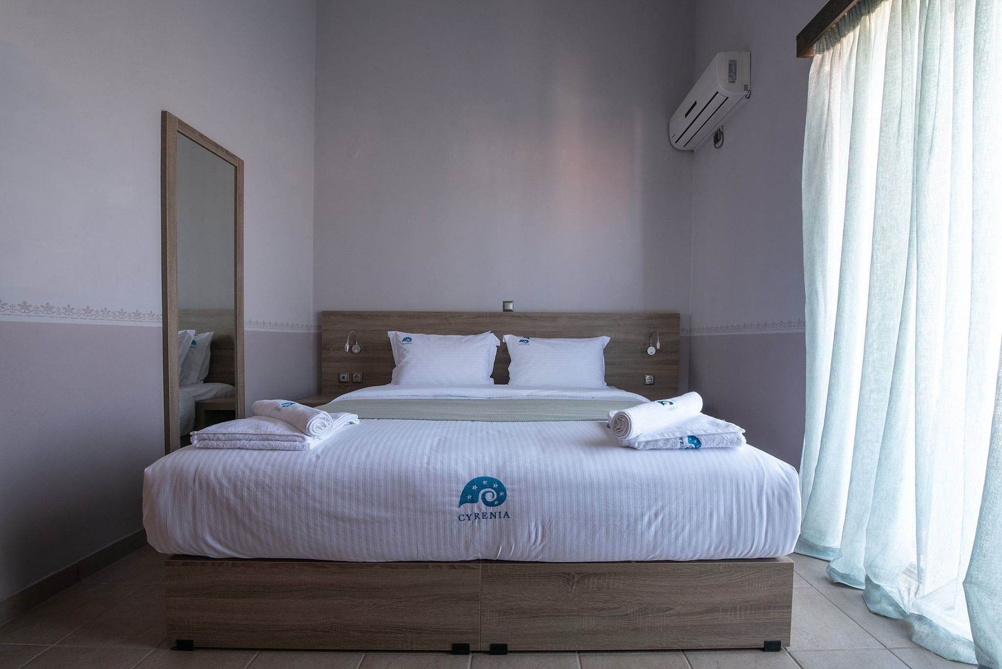 Een net opgemaakt bed met een logo, een houten hoofdbord, witte beddengoed en een nachtkastje met een lamp.
