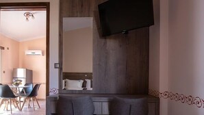 Habitación de hotel con cabecera de madera, televisor de pantalla plana en la pared, dos sillas grises y una mesita pequeña.