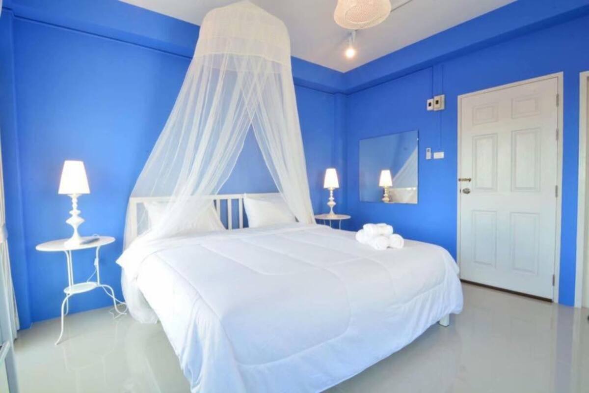 A bedroom with a bed, white bedding, a white bedside table, a white lamp, a white door, and a white ceiling fan.