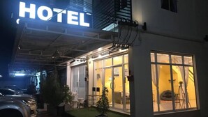 Entrada de un hotel con un cartel, un coche aparcado fuera y una pequeña zona de estar al aire libre.