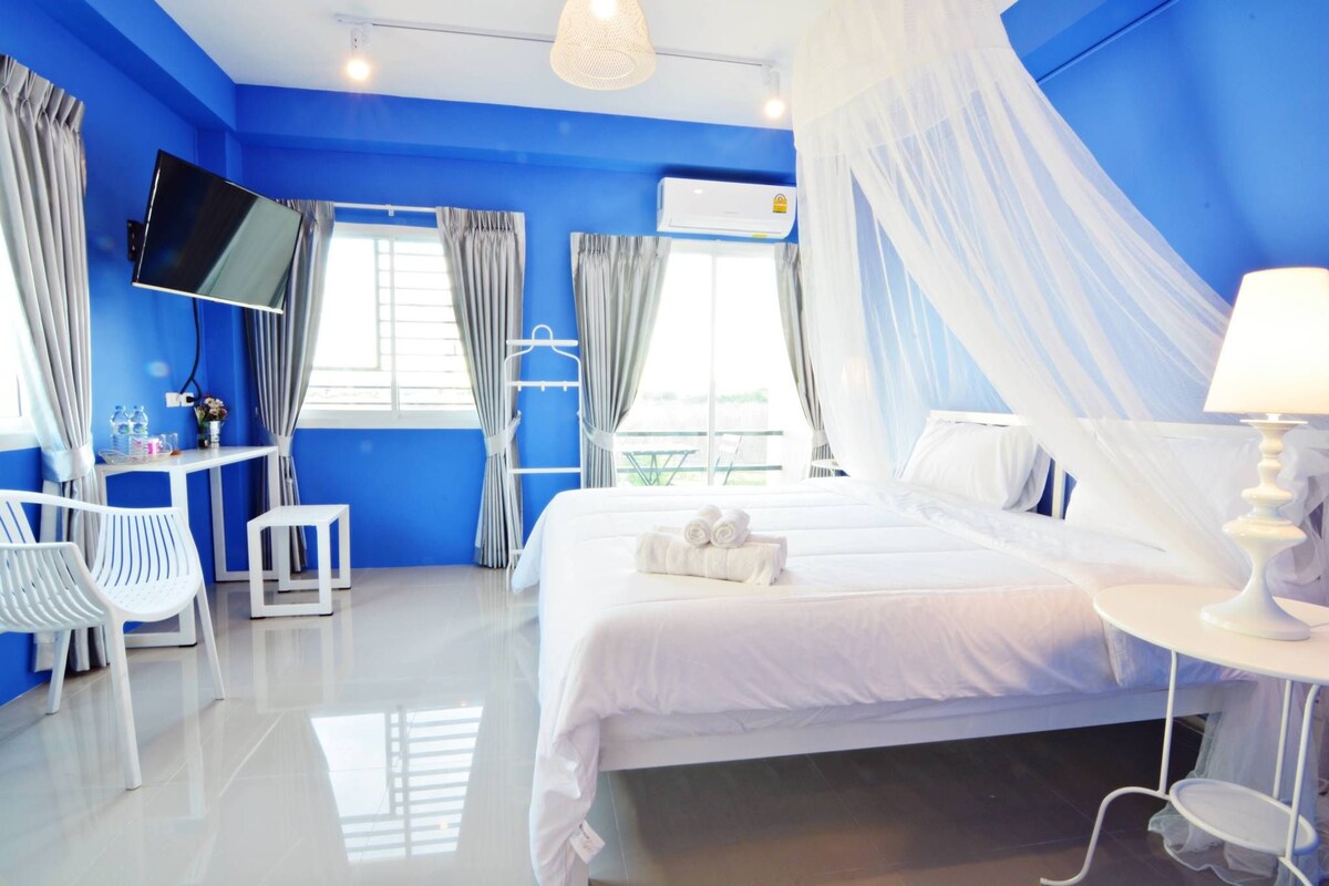 A hotel room with a large bed, a TV mounted on the wall, a desk with a chair, and a window with curtains.