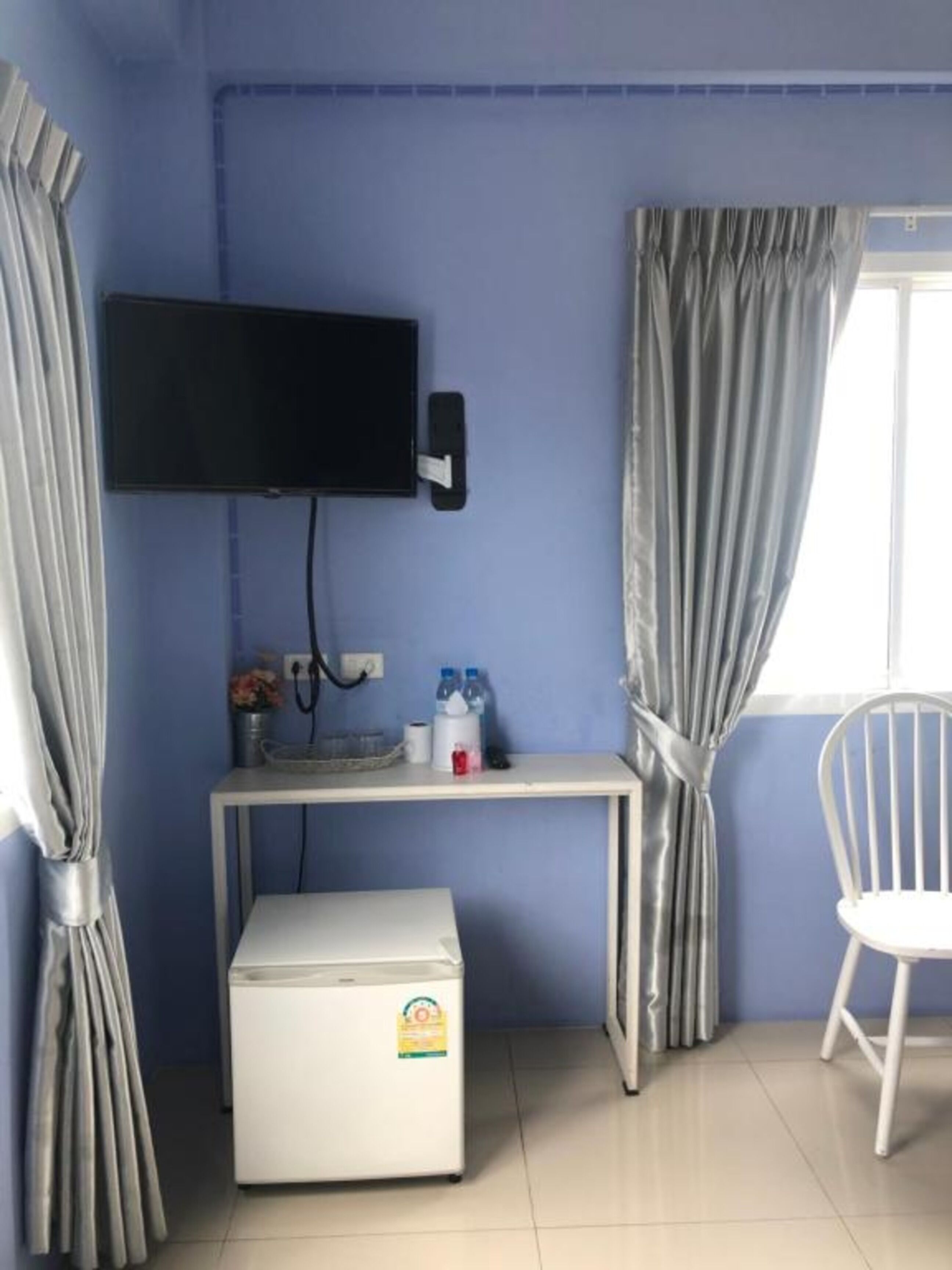 Une pièce avec un mur bleu, un téléviseur à écran plat fixé au-dessus d’un bureau blanc, un petit réfrigérateur et une chaise blanche.