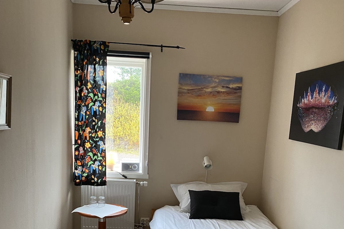 Ein Schlafzimmer mit Bett, kleinem Tisch, Sessel, Fenster mit Vorhängen, Heizkörper und einem Bild an der Wand.