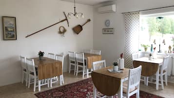 A dining area with white chairs and wooden tables, a red patterned rug, a chandelier, and a window with curtains.